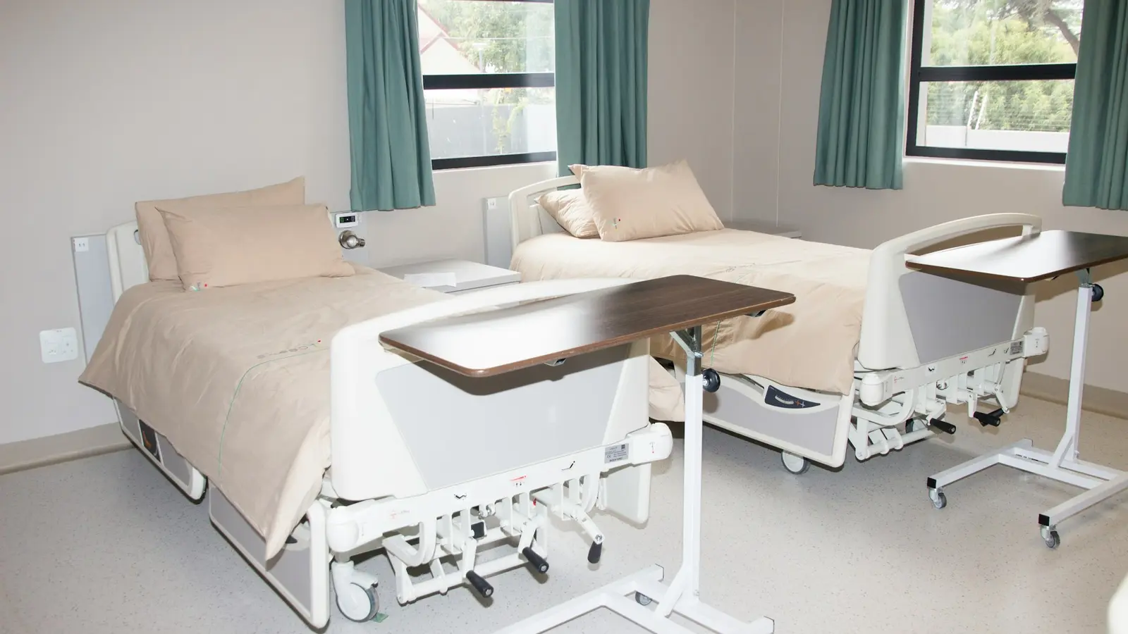 Two made hospital beds with beige linen and wheeled overbed tables in an empty ward room with teal curtains at the windows