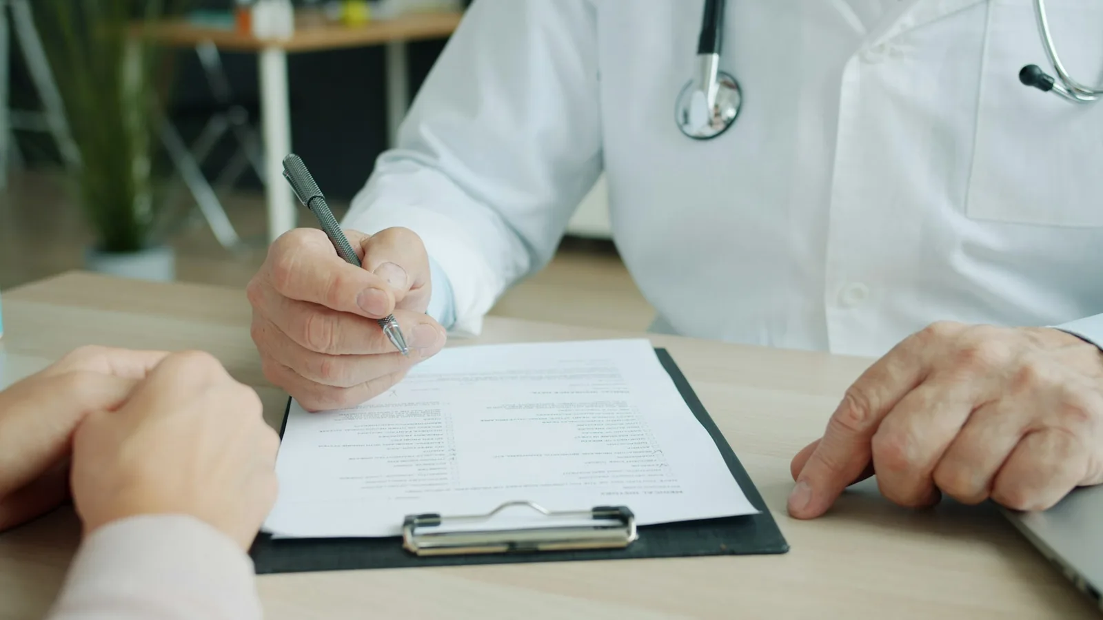 A doctor in a white coat writes on a clipboard form across the desk from a patient