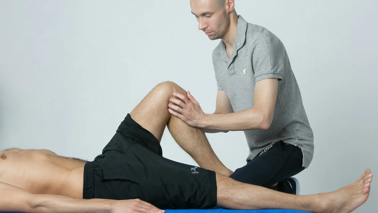 An Australian allied health treatment room in afternoon light, a physiotherapist's hands steadying a participant at the edge of a plinth partway through a transfer